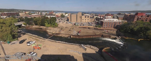 Aerial view of the Union Street Dam FishPass construction site in Traverse City, Michigan. The image shows a partially completed structure spanning the Boardman River, with visible sheet piling, construction equipment, work vehicles, and surrounding urban buildings under clear skies.