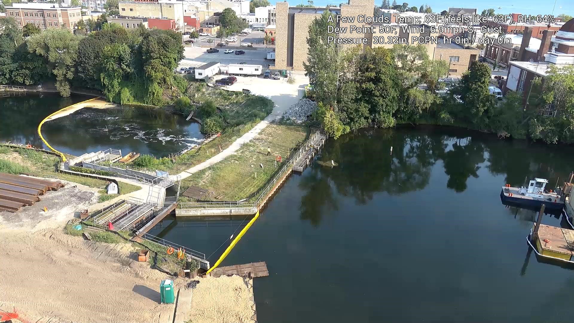 Aerial view of a construction site by a river, showing a containment boom across the water, construction materials and equipment along the shoreline, and a partially built structure near the water's edge. In the background, there are trees, buildings, and a parking area. A small boat is docked near the right edge of the water. The image includes weather information in the top right corner, reporting a temperature of 63°F, wind speed of 6.9 mph, and a few clouds in the sky.